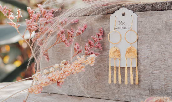 Nic Danning Jewellery profile banner. A pair of sparkling hammered brass earrings hang from a putty grey swing tag amongst pretty pastel dried flowers.