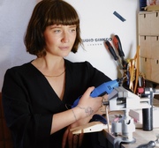 Girl with brown bob sat at workbench holding a dremel, smiling and looking off to her left hand side  