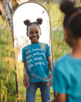 A female child wearing a Ocean Tur-Shirt that reads "I am brave. I am strong. I am kind. I am loved" whilst looking in the mirror.