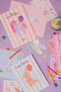 A coloured in unicorn on a personalised birthday card, with a sticker of a unicorn to the side, and a dinosaur happy birthday card with a cupcake