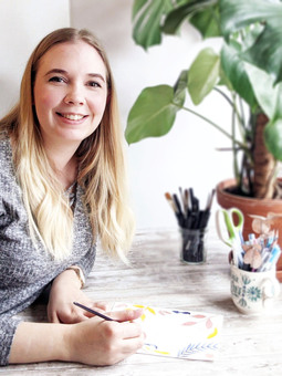 Eva sits painting at a table; behind her are a plant and two pots of pens.