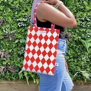 Black woman wearing blue jeans and black vest top carrying a red and white diamond print bag over the shoulder in front of green plants outside..