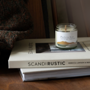 earth and vine wooden wicked petrichor candle on top of two cream coloured home decor books on a wooden mid century chair draped with a brown speckled scarf