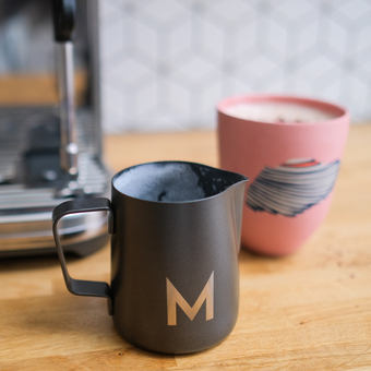 A black milk jug with the initial M engraved on it and in the background a pink cup with hot chocolate in it and part of a coffee machine