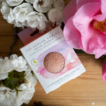 A pink konjac sponge presented in pretty pink box with pink and purple design, sat on a wooden table amongst summery flowers