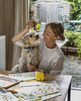 An image of a blonde girl and her dog sitting at a table painting in watercolour