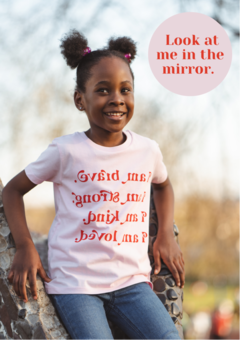 A female child wearing a pink & red Tur-Shirt that reads "I am brave. I am strong. I am kind. I am loved" backwards. Sticker reads "Look at me in the mirror.