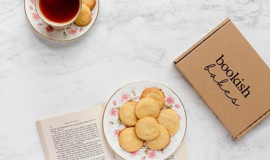 A bookish bakes baking kit sits on a marble countertop beside an open book topped with a plate of lemon biscuits and a cup of tea.