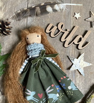 Wildflower Fairy with green illustrated dress, blue scarf and London’s brown hair, laying on a wooden background.