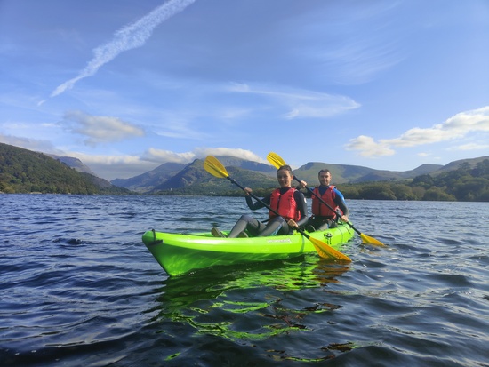 A couple paddles together in a green sit-on-top kayak on Llyn Padarn Lake, with the majestic Mount Snowdon towering in the background under a clear blue sky. Th