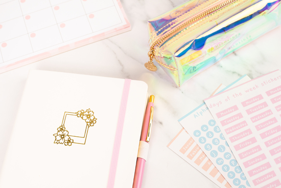 An image of sakura polaroid white bullet journal, clear holographic pencil case, sticker sheets and a monthly planner