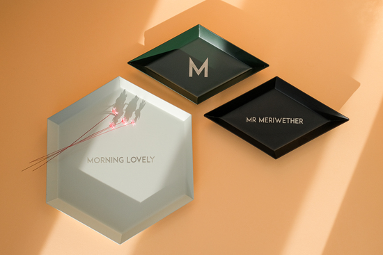 A pale orange background with three geometric trays in the foreground, each engraved with a different message; Morning Lovely, Mr Meriwether and M