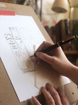 Georgina's hand holding a black fountain pen sketches a book spine onto a piece of paper mounted on an easel, a shelf of books can be seen in the background