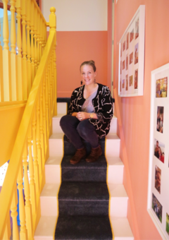 A white woman with brown hair sits on her stairs. The walls are coral pink and the bannisters are painted bright yellow.