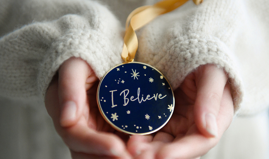 A navy blue Christmas decoration with the words "I Believe" in gold lettering is being held in two hands.