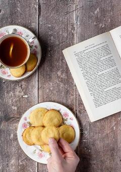 A hand reaches for a lemon biscuit on a rose patterned plate next to an open book and a cup of tea.