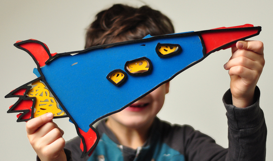 a boy is holding in front of his face a laser cut out of his drawing of a rocket. The rocket is blue with red and yellow detail.