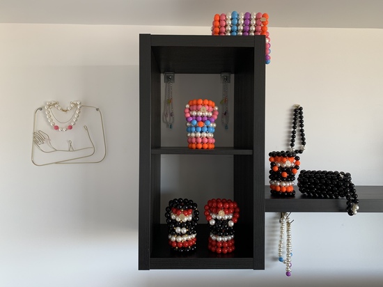 Six beaded buckets bags in different colour combinations arranged on three shelves. There are beaded necklaces hanging from the wall