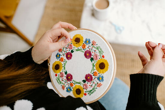 A birds eye view of a woman's hands embroidering a wreath made up of sunflowers and other summer flowers. A cup of tea sits on a table in the background.