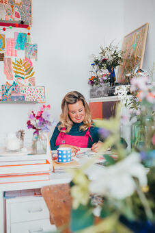 Floradore artist Lucy Innes Williams at work in her studio. Woman sat at desk surrounded by flowers in a studio.