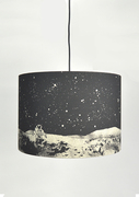 A large drum lampshade hanging on a black electrical wire. The lampshade is black and grey, with an image of an astronaut on the moon on it.