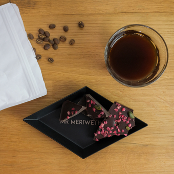 A wooden background with coffee beans and a cup of coffee. A black diamond shaped tray has chocolate chunks on it with the words Mr Meriwether engraved on it