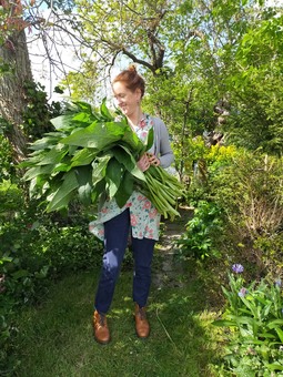 A woman stands with an armful of comfrey in a garden with shrubs and trees
