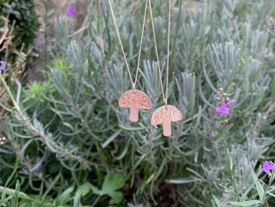 Two 9ct gold mushroom necklaces placed on a background of a rosemary plant