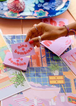 Brown woman's hand holding business card that says "Nina Bombina" against a background of a pile of art prints