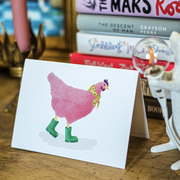 A stylised watercolour illustration on a greeting card of a pink chicken, wearing green wellies and a yellow scarf, against a background of white and pink books