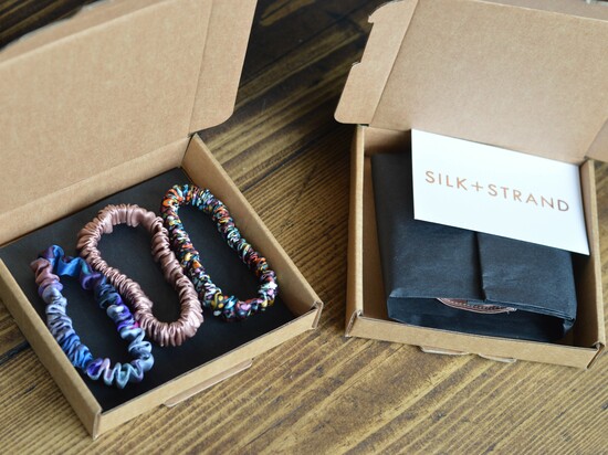 A variety of skinny silk hair ties in a cardboard postal box and next to it another box with a silk scrunchie wrapped with black tissue paper and foil card